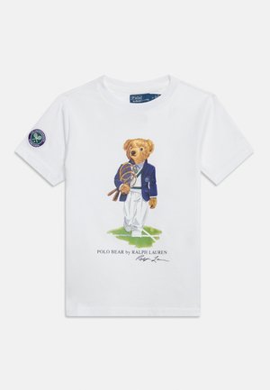 Maglietta bianca con un orsacchiotto vestito con abbigliamento da tennis che tiene le racchette, in piedi su un campo da tennis verde con una toppa Wimbledon sulla manica.