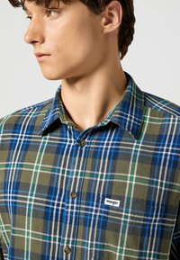 Camicia in flanella a quadri verde e blu con cuciture bianche e etichetta Wrangler. Presenta un colletto e chiusura frontale con bottoni. Tessuto testurizzato.