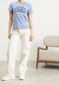 T-shirt di cotone azzurro chiaro con testo "Original Gant Sportswear", abbinato a pantaloni a gamba larga color avorio e sneakers bianche.