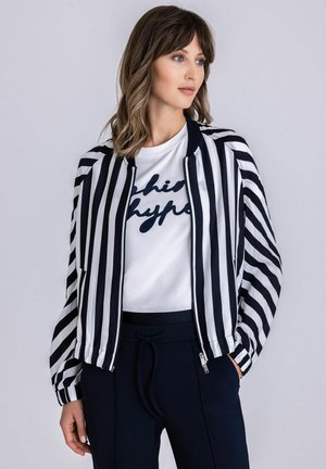 Frau mit schulterlangem Haar, die eine schwarz-weiß vertikal gestreifte Jacke, ein weißes T-Shirt mit Grafik und dunkle Hosen trägt und seitlich blickt.