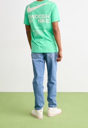 Osoba stojąca tyłem, ubrana w zieloną koszulkę Nike z białym napisem i logo, jasnoniebieskie jeansy oraz białe trampki na zielonej podłodze.