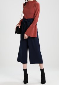 Rostfarbener, gerippter Langarmshirt mit Glockenärmeln, kombiniert mit hochgeschnittenen, marineblauen Culottes. Das Model ergänzt das Outfit mit einer schwarzen Handtasche und Ankle Boots.