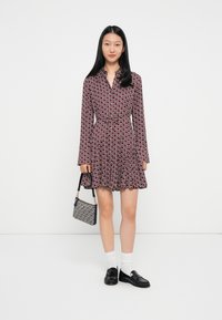 Sarkans un balts rakstains kleitas modelis ar garajām piedurknēm, apkakli un jostu viduklī. Modelis tur nelielu rakstainu rokassomu ar melnu siksniņu.