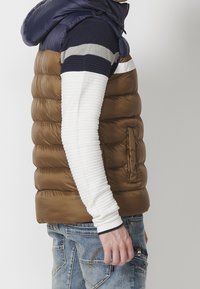 Brown puffer vest with navy hood, side pockets, and ribbed white long-sleeve shirt underneath. Worn with blue denim jeans.