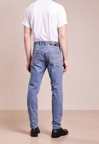 Person som bär ljusblå jeans, vit t-shirt med kort ärm och svarta skor, står vänd bort från kameran mot en beige bakgrund.