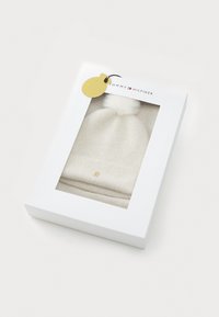Witte gebreide beanie met een pompon, voorzien van een gemêleerd patroon, omgeslagen boord en gouden logo, gepresenteerd in een heldere geschenkdoos.