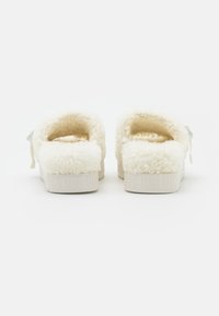 Zapatillas de shearling blancas con una textura suave, forma redondeada en la parte trasera y suelas de goma con el nombre de la marca en relieve. Acentuadas con una etiqueta azul.