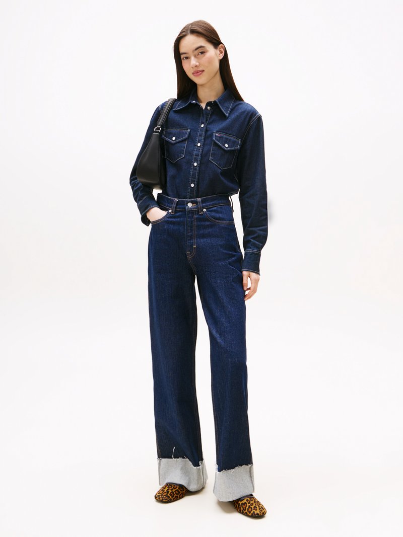 Camicia in denim di colore blu scuro, maniche lunghe e due tasche frontali. Abbinata a jeans a gamba ampia blu scuro con orli grigi sfilacciati.