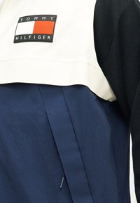 Έμβλημα με το λογότυπο Tommy Hilfiger σε λευκό και μαύρο μπουφάν φορεμένο πάνω από ένδυμα σε σκούρο μπλε χρώμα.