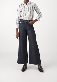 Frau, die eine weite dunkle Jeanshose, ein gestreiftes weißes Hemd mit Blumenmuster und schwarze Absatzmules trägt, dabei mit einer Hand in der Tasche steht.