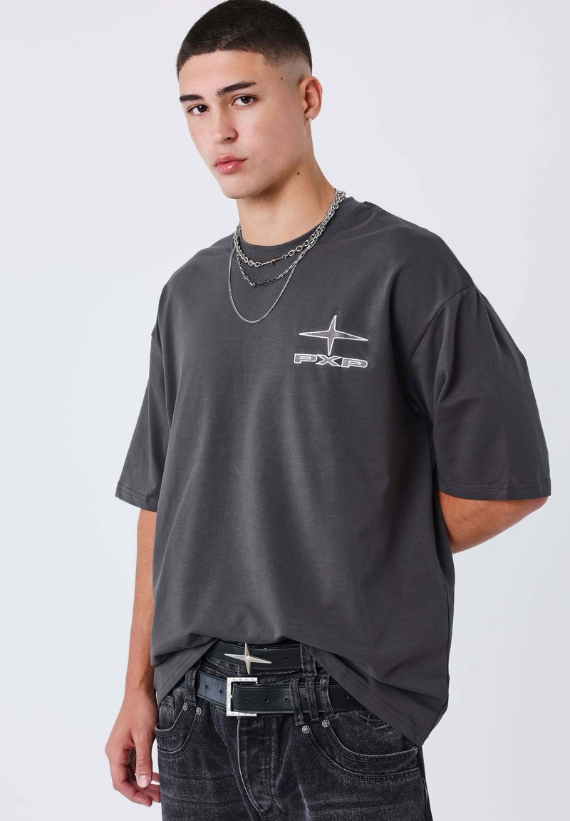 Camiseta gris de manga corta con un logo en la parte frontal, combinada con unos pantalones vaqueros negros texturizados y un cinturón negro con una hebilla plateada. Cadenas visibles alrededor del cuello.