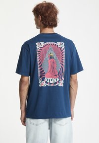 Ung mand med krøllet hår iført en marineblå T-shirt, der har en pink abstrakt knælende figur og ordet "STONE" på ryggen.