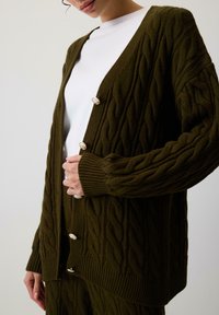 Tamsiai žalia megzta kardiganas su pynimo raštu, V formos iškirpte, ilgomis rankovėmis ir dekoratyviais mygtukais priekyje.