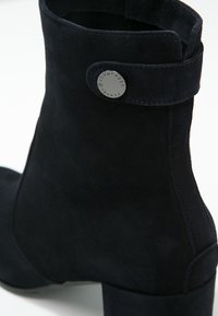 Bottine en daim noir avec talon bas à bloc et petit bouton métallique rond gravé du nom de la marque sur la lanière latérale.