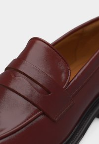 Λείο γυαλισμένο καστανόσκούρο δερμάτινο μοκασίνι με λουράκι πέννι και μαύρη καουτσούκ σόλα, με εμφανείς λεπτομερείς ραφές και εσωτερική επένδυση.