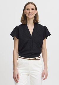 Blusa nera a maniche corte con scollatura a V, caratterizzata da pattern a pois in rilievo. Abbinata a pantaloni bianchi e una cintura marrone.