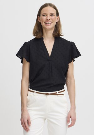 Must have lühikeste varrukatega must Blouse, millel on V-kaelus ja tekstuursete täppidega muster. Kombineeritud valgete pükstega ja pruuni vööga.