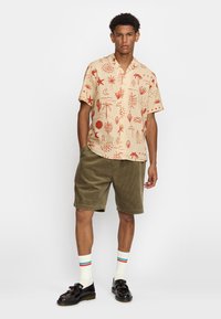 Korte mouw beige shirt met rode geometrische en botanische prints, gecombineerd met olijfgroene korduroy shorts en witte sokken met kleurstripes.