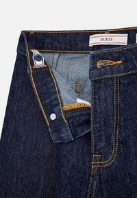 Dunkelblaue Jeans aus Denim mit gelben Nähten, einem Knopfverschluss und einem Etikett mit roten Akzenten. Das Innenfutter hat hellere Stoffdetails.