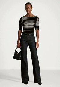 Γυναίκα με fitted τοπ σε μαύρες και κίτρινες ρίγες με μανίκια έως τον αγκώνα, υψηλή μαύρη παντελόνα με φαρδιές leg, κρατώντας μια μικρή μαύρη τσάντα χεριού.