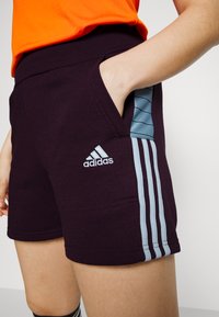 Dunkelviolette Sportshorts mit hohem Bund, hellblauen Seitenstreifen und einem gestickten weißen Adidas-Logo auf der Vorderseite.