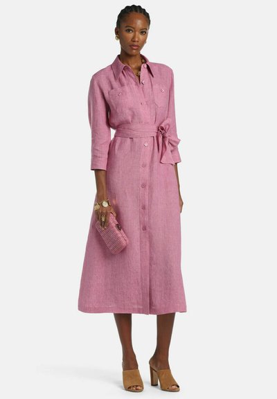 Frau trägt ein mittellanges rosa Kleid mit Knopfleiste, 3/4-Ärmeln, gebundener Taille, hält eine passende rosa Clutch und trägt beige offene Mules mit Absatz.