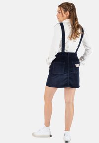 Marineblauwe fluwelen jurk met verstelbare schouderbanden, met twee achterzakken en een lichte A-lijnvorm. G gedragen met witte sneakers.