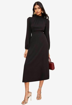 Schwarzes Midi-Kleid mit langen Puffärmeln, hohem Kragen und taillierter Taille. Kombiniert mit nude-farbenen Strappy-Heels und einer kleinen bordeauxfarbenen Handtasche.