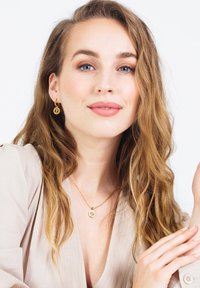 Gouden ketting en oorringen hebben ronde hangers met een gegraveerde letter. Model heeft golvend haar dat over een lichte blouse valt.