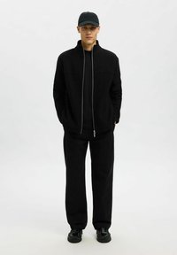 Svart fleecejacka med dragkedja och hög krage, bärs över en svart t-shirt, matchad med lösa svarta byxor och svarta skor; baseballkeps.