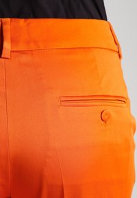 Pantaloni arancioni con una texture liscia, caratterizzati da una tasca posteriore con chiusura a bottone e cuciture a contrasto lungo la cucitura della tasca.
