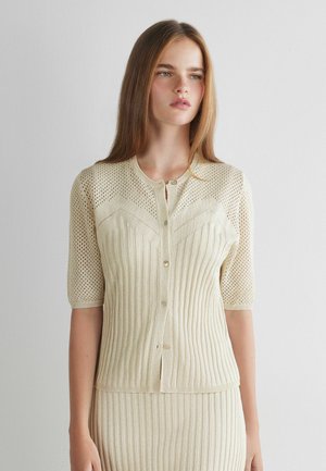 Junge Frau mit langen braunen Haaren, die ein beiges, kurzärmliges, geknöpftes Strickoberteil und einen passenden, gerippten Rock vor einem schlichten Hintergrund trägt.