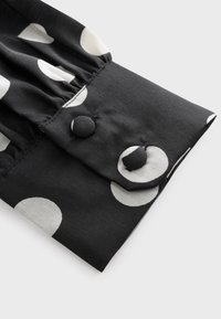 Poignet en tissu noir avec de gros pois blancs, comprenant deux boutons recouverts de tissu et une couture froncée près du poignet.