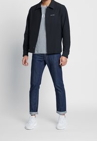 Sort skjortejakke over en grå t-shirt, parret med mørkeblå jeans. Hvide sneakers fuldender looket, som har opkrøllede ærmer.