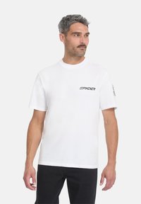 Hombre con cabello entrecano que lleva una camiseta blanca de Spyder y pantalones negros, mirando ligeramente hacia su derecha contra un fondo liso.