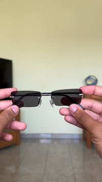 Óculos de sol retangulares pretos com acabamentos metálicos, hastes finas e almofadas nasais transparentes, segurados com duas mãos contra um fundo neutro.