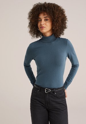 Un pull col roulé bleu en tricot, doté de longues manches et d'une silhouette ajustée, associé à un jean noir et une ceinture.