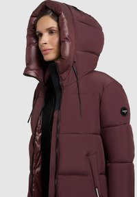 Jachetă puffer burgundă cu guler înalt, glugă mare și textură lucioasă. Prezintă un logo negru și un fermoar negru, precum și buzunare laterale.