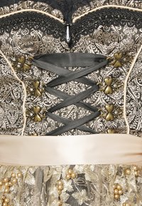 Gros plan sur un corset orné avec un ruban noir à lacets, tissu brocart floral doré, fermoirs métalliques en forme de cœur et partie jupe en dentelle ornée de perles.