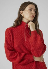 Maglione rosso lavorato a maglia con colletto alto e chiusura con zip. Presenta una texture a coste diagonali e maniche lunghe.