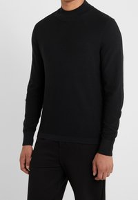 Homme portant un pull à col roulé noir ajusté à manches longues et un pantalon noir, debout devant un fond gris clair uni.