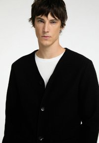 Mustmust kaartigan V-kaelusega, varrukatega ja nupuga sulgemisega, pehme tekstuuriga. Kandmine valge T-särgiga toob välja kontrasti.