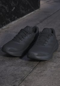 Двойка черни маратонки Adidas с връзки, поставени на сив плочкиран под близо до тъмен стенен ъгъл.