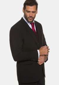 Costume noir bien ajusté, avec un revers cranté, une fermeture à trois boutons et une texture subtile. Accentué par une cravate rouge et une chemise blanche.