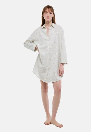 Femme debout pieds nus portant une robe-chemise ample à manches longues blanche à motif floral sur un fond uni.