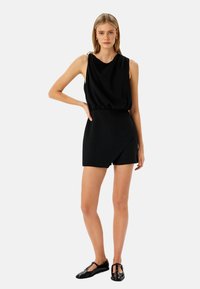 Vestido negro sin mangas con escote drapeado y dobladillo asimétrico, hecho de una tela suave. Combinado con planos de charol negros.
