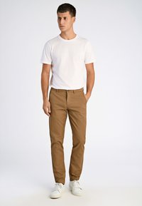 Bruna bomulls-chinos med slim fit, bärs med en vit t-shirt med kort ärm och vita sneakers, stående mot en neutral bakgrund.