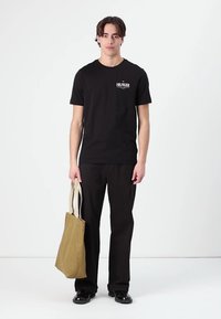 Zwarte katoenen T-shirt met een bedrukte logo, gecombineerd met zwarte broeken. Het model houdt een beige totebag vast met dubbele handvatten.