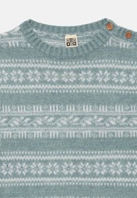 Lys blå strikket sweater med ribbet rund hals, prydet med hvide geometriske mønstre og to træknapdetaljer på skulderen.