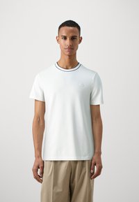 T-shirt bianco a maniche corte con colletto a coste, caratterizzato da una striscia blu navy a contrasto e un logo ricamato sul lato sinistro del petto.